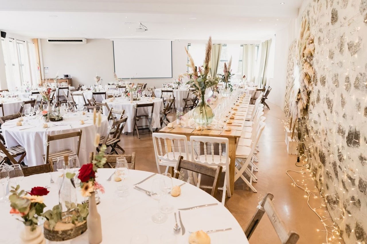 Salle de réception lumineuse avec tables décorées de fleurs, chaises en bois et murs en pierre apparente.