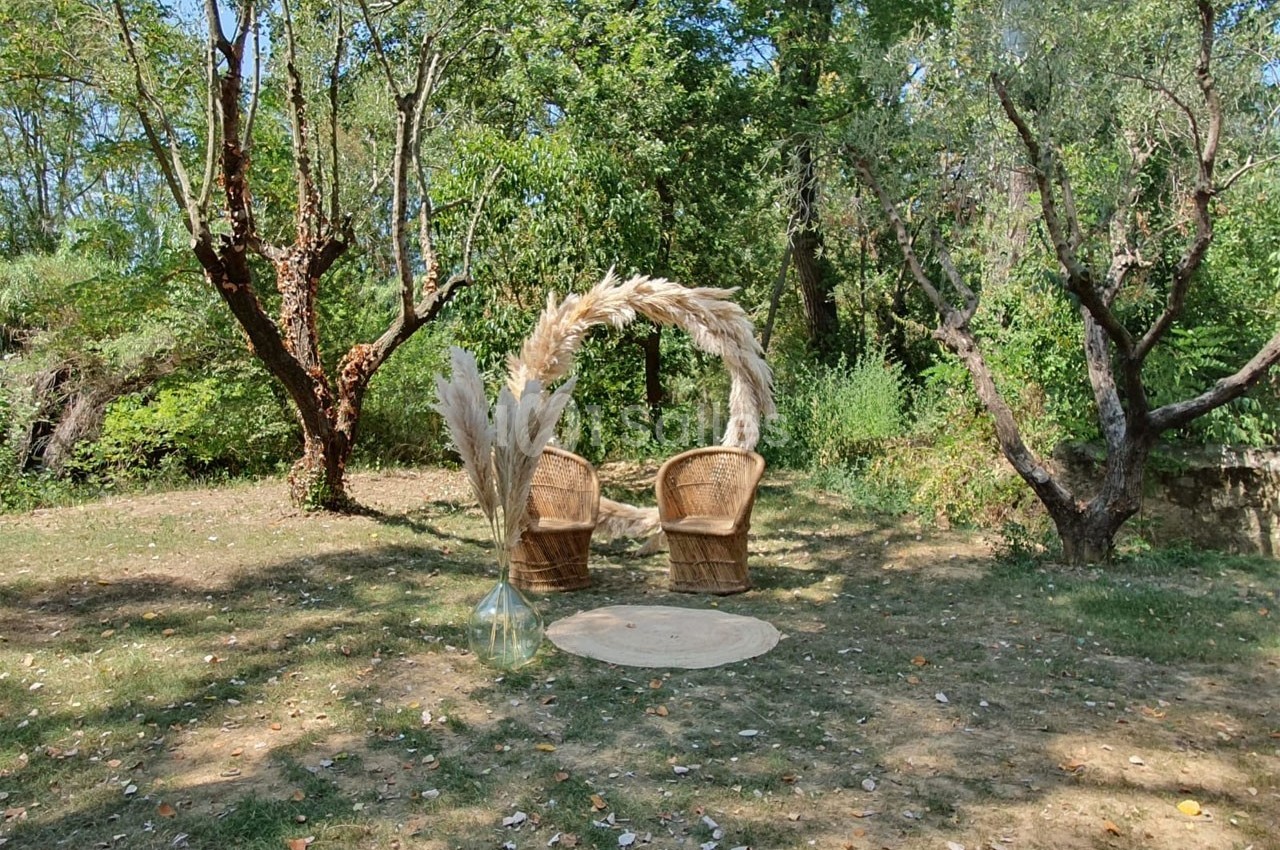 Deux fauteuils en rotin disposés sous une arche décorée de pampas, dans un jardin arboré.