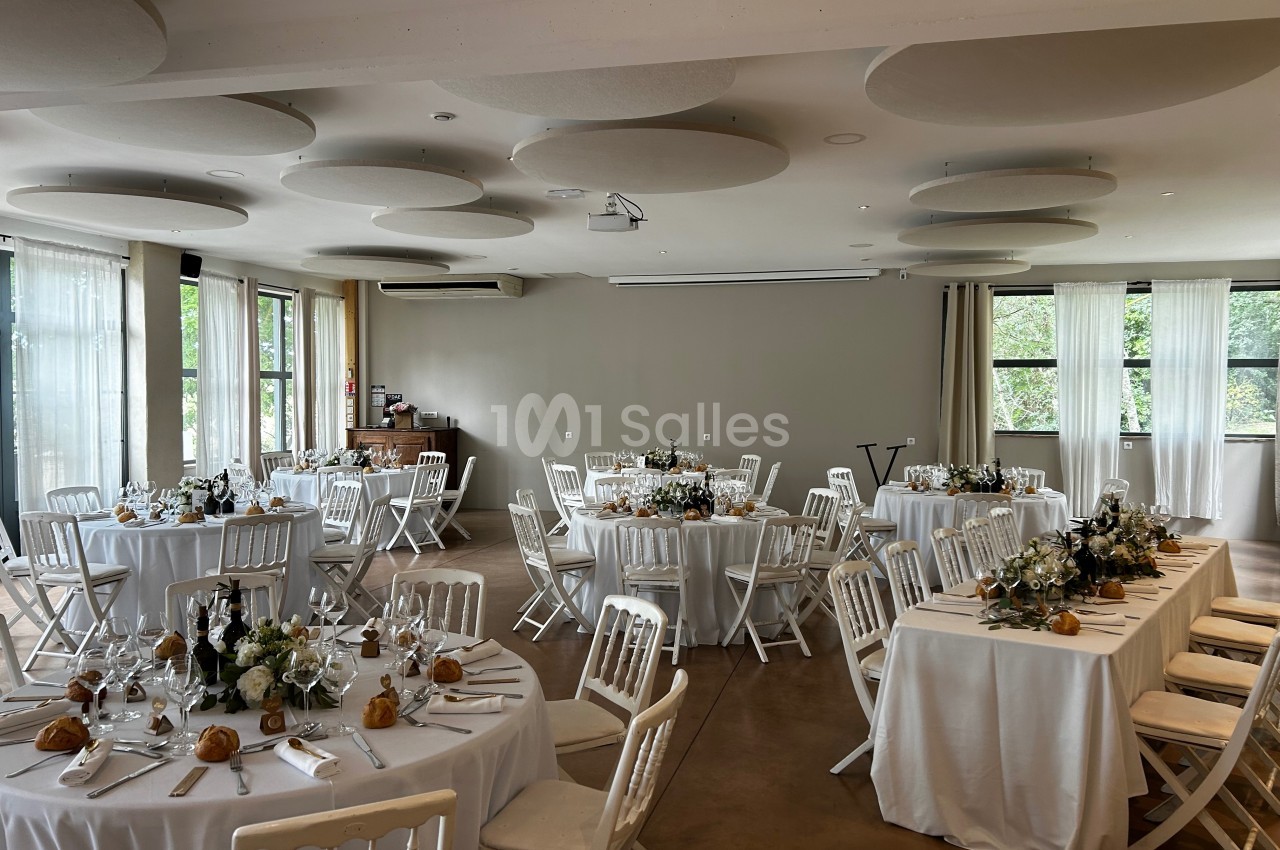 Salle de réception lumineuse avec des tables rondes décorées de fleurs et dressées pour un repas.