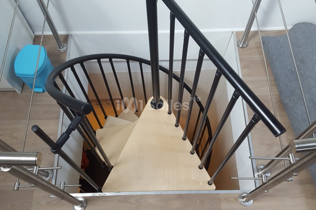 Escalier en colimaçon avec marches en bois clair et garde-corps en métal noir, vu depuis le haut.