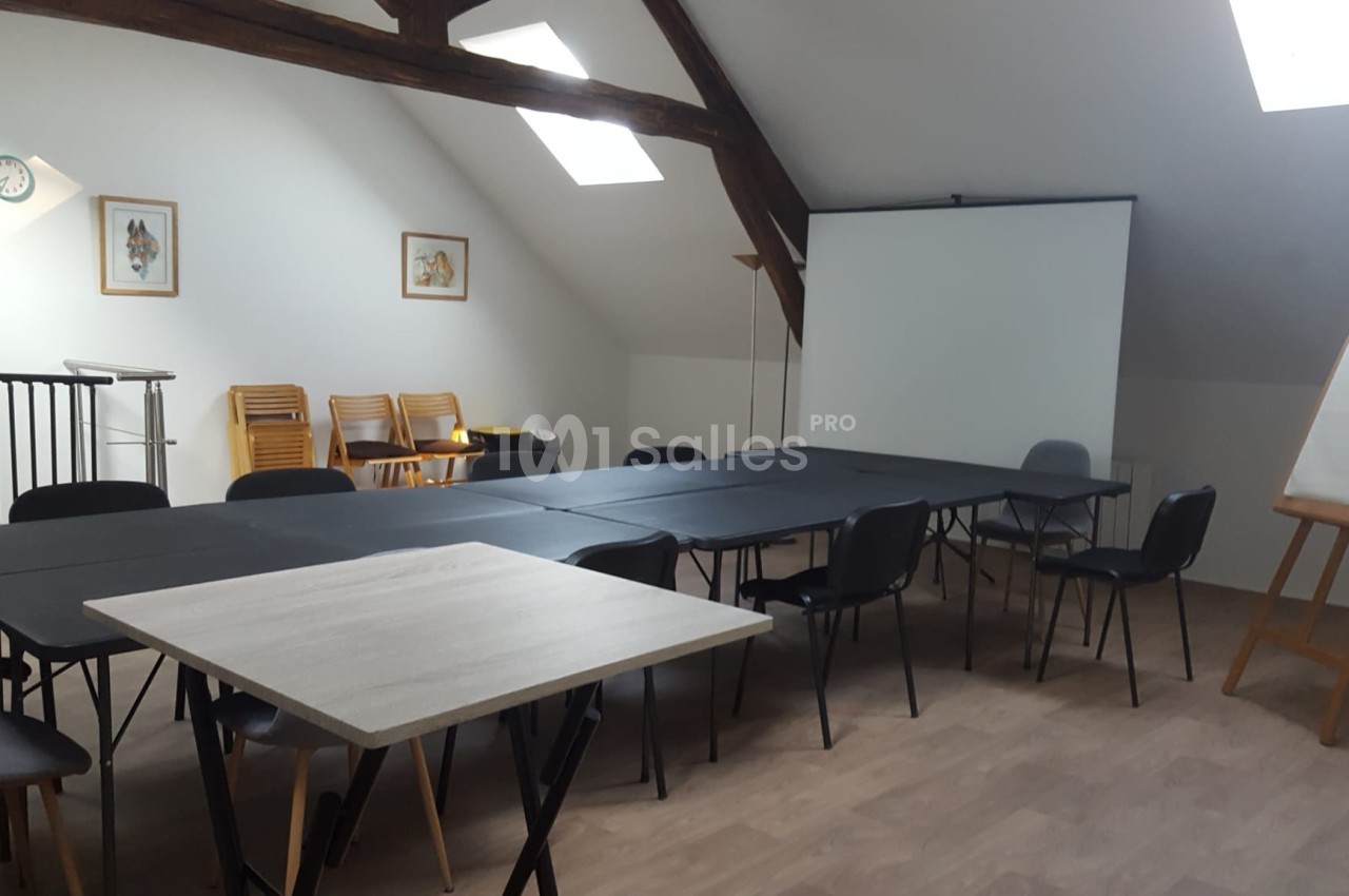 Salle de réunion avec tables noires, chaises, tableau blanc sur chevalet et écran de projection sous un toit mansardé.
