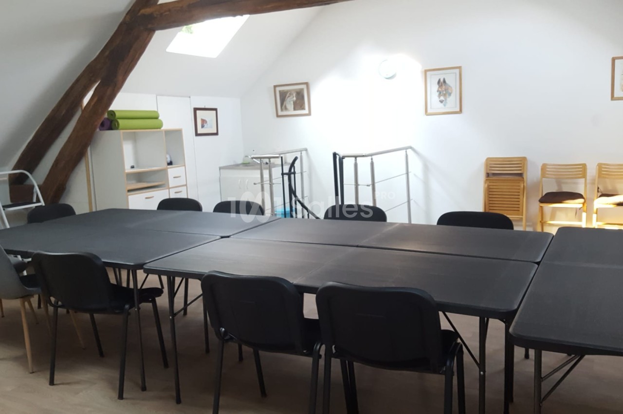 Salle lumineuse avec tables noires disposées en rectangle, chaises autour, étagères et cadres accrochés aux murs blancs.