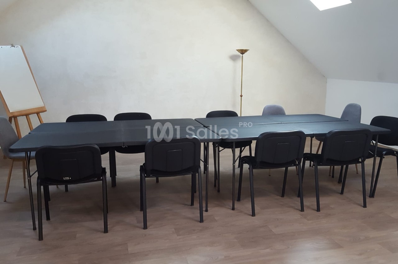Salle de réunion avec deux grandes tables noires, plusieurs chaises, un tableau chevalet et un lampadaire.