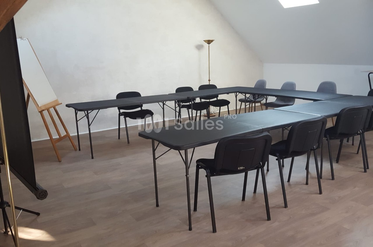 Salle de réunion lumineuse avec tables noires disposées en U, chaises noires et grises, tableau chevalet et lampadaire.