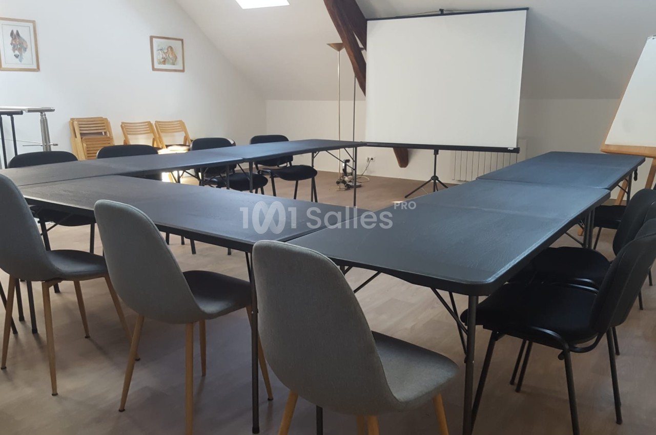 Salle de réunion lumineuse avec tables disposées en U, chaises, tableau blanc et écran de projection.