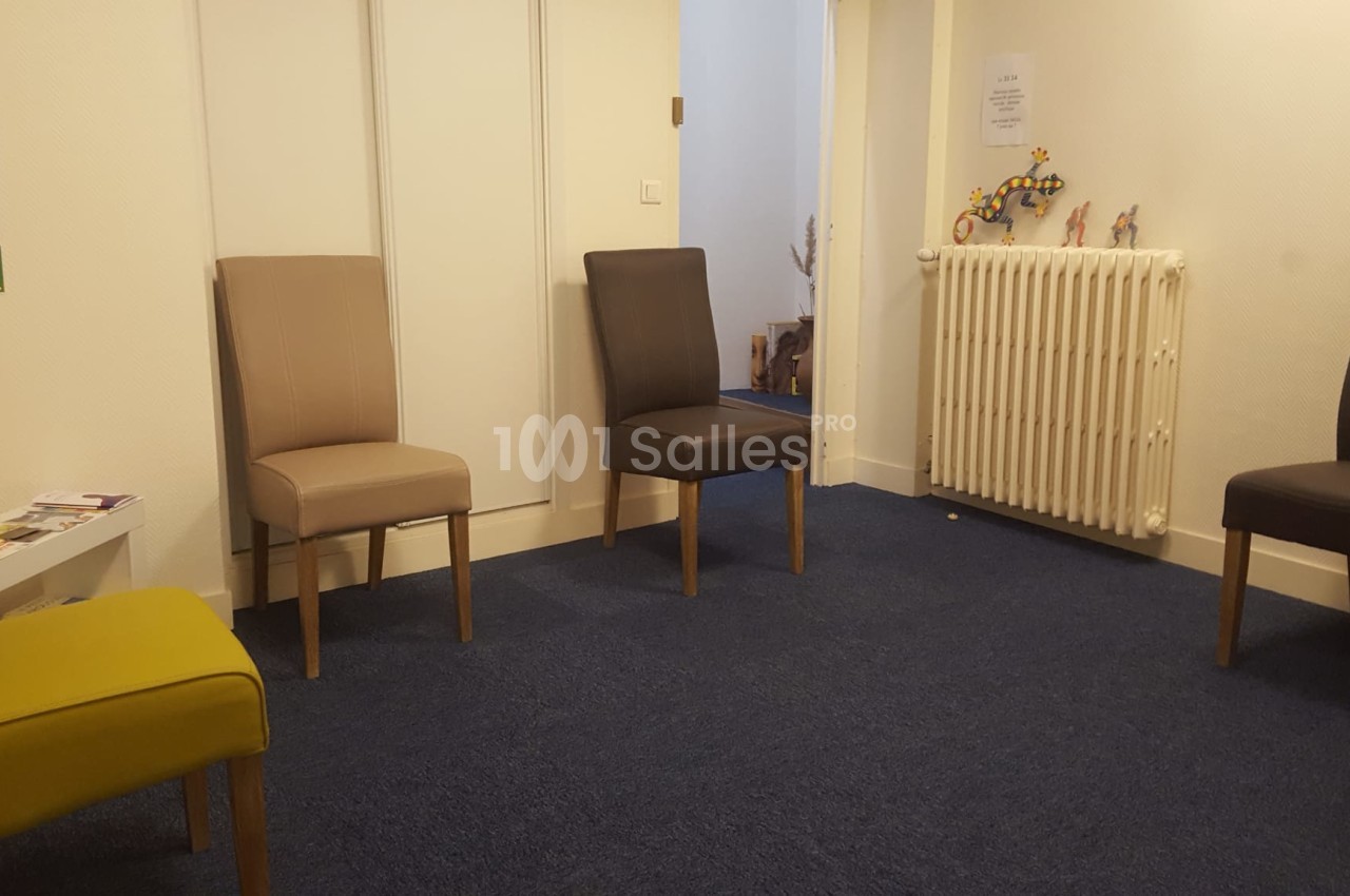 Salle d'attente avec quatre chaises disposées autour d'un radiateur, sol en moquette bleue et murs clairs.
