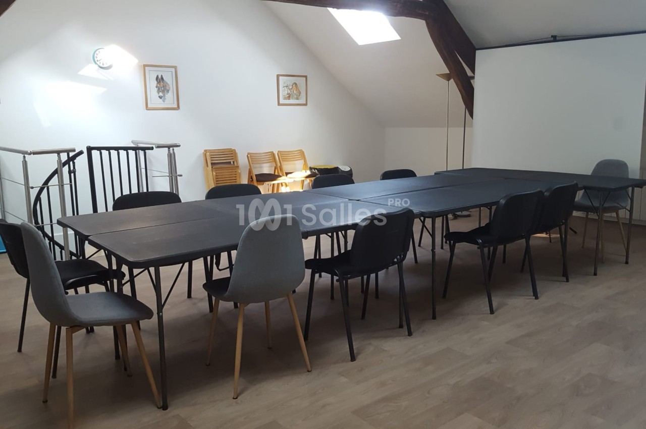 Salle de réunion lumineuse avec tables noires alignées, chaises variées et écran de projection en arrière-plan.