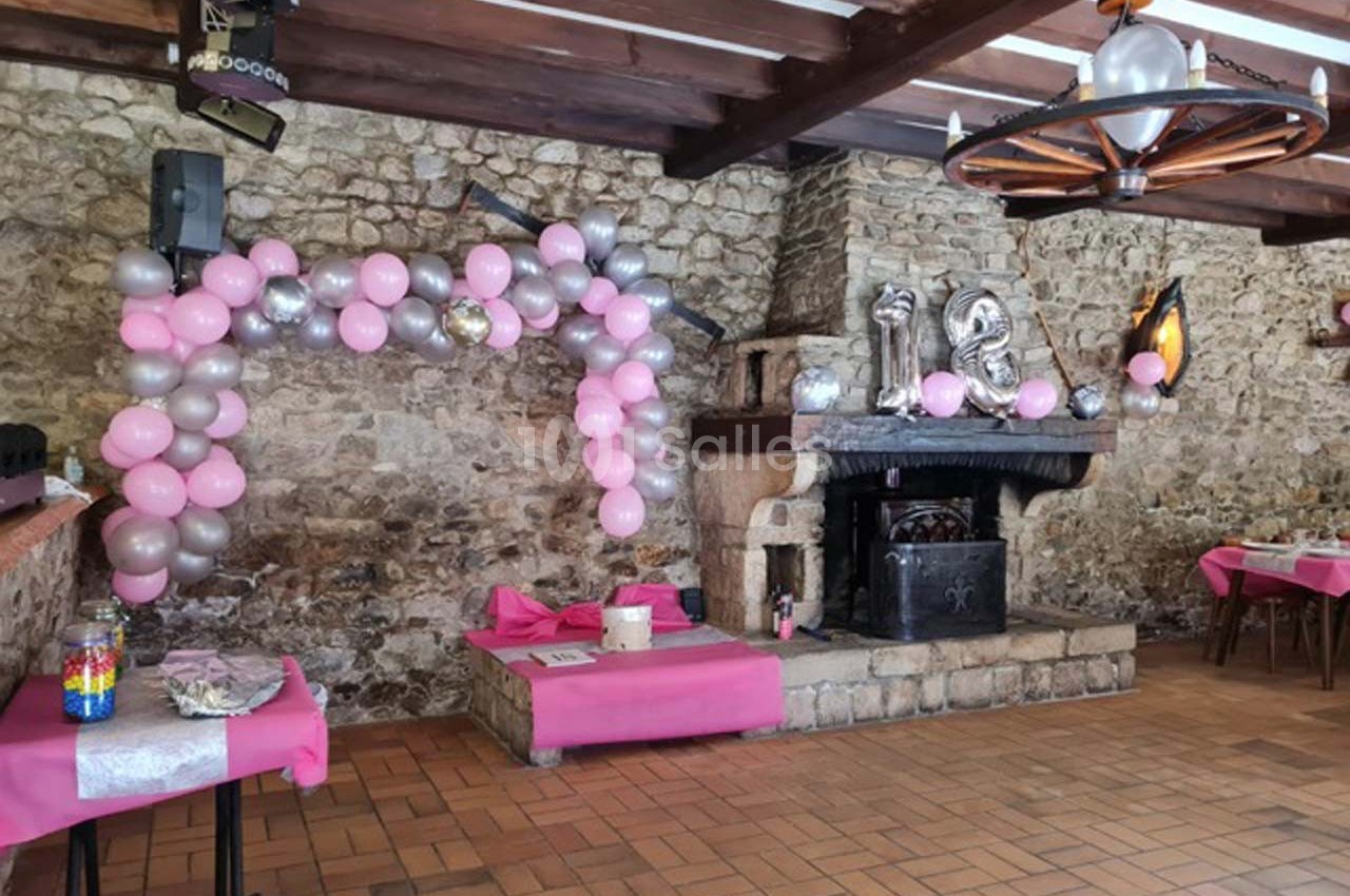 Salle décorée pour une fête, avec des ballons roses et argentés près d'une cheminée en pierre.