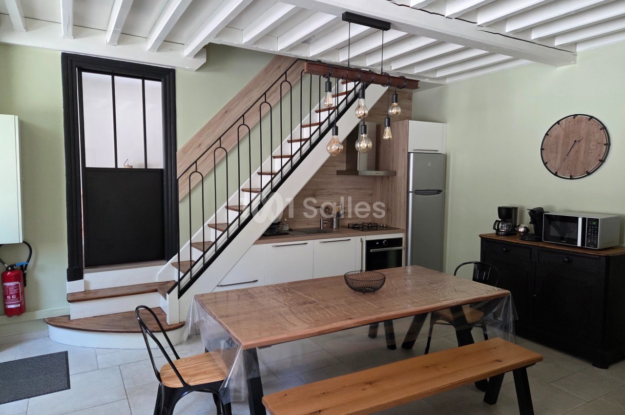Cuisine moderne avec table en bois, chaises métalliques, escalier en bois, et électroménagers sous un plafond à poutres…