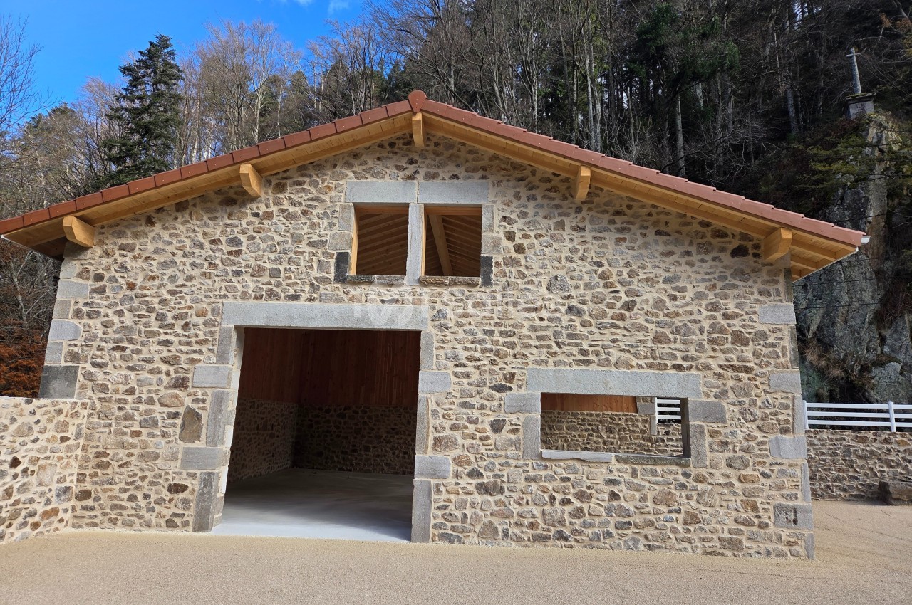 Bâtiment en pierre avec toit en tuiles, ouvertures rectangulaires, situé devant une forêt.