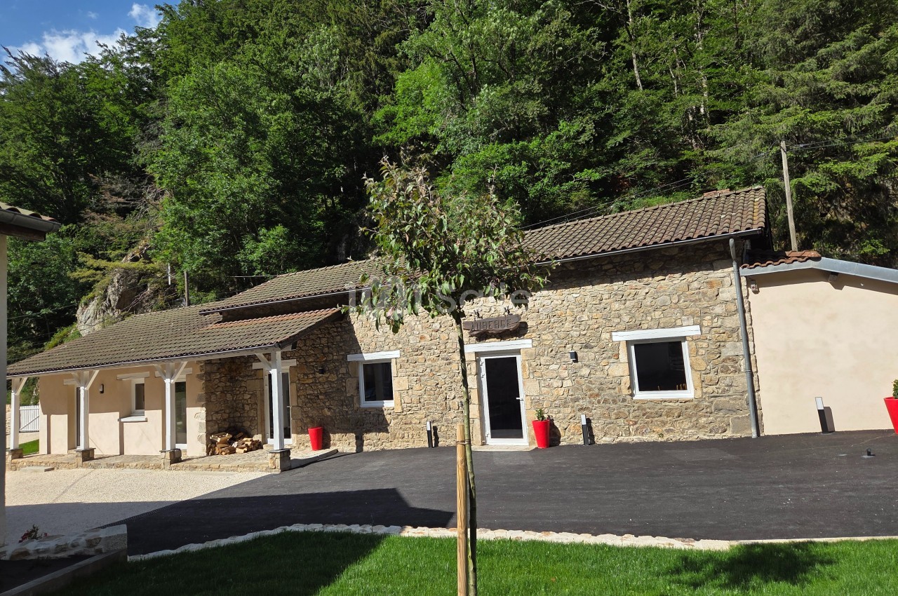 Façade d'une maison en pierre avec toit en tuiles, entourée de verdure et d'une allée asphaltée.