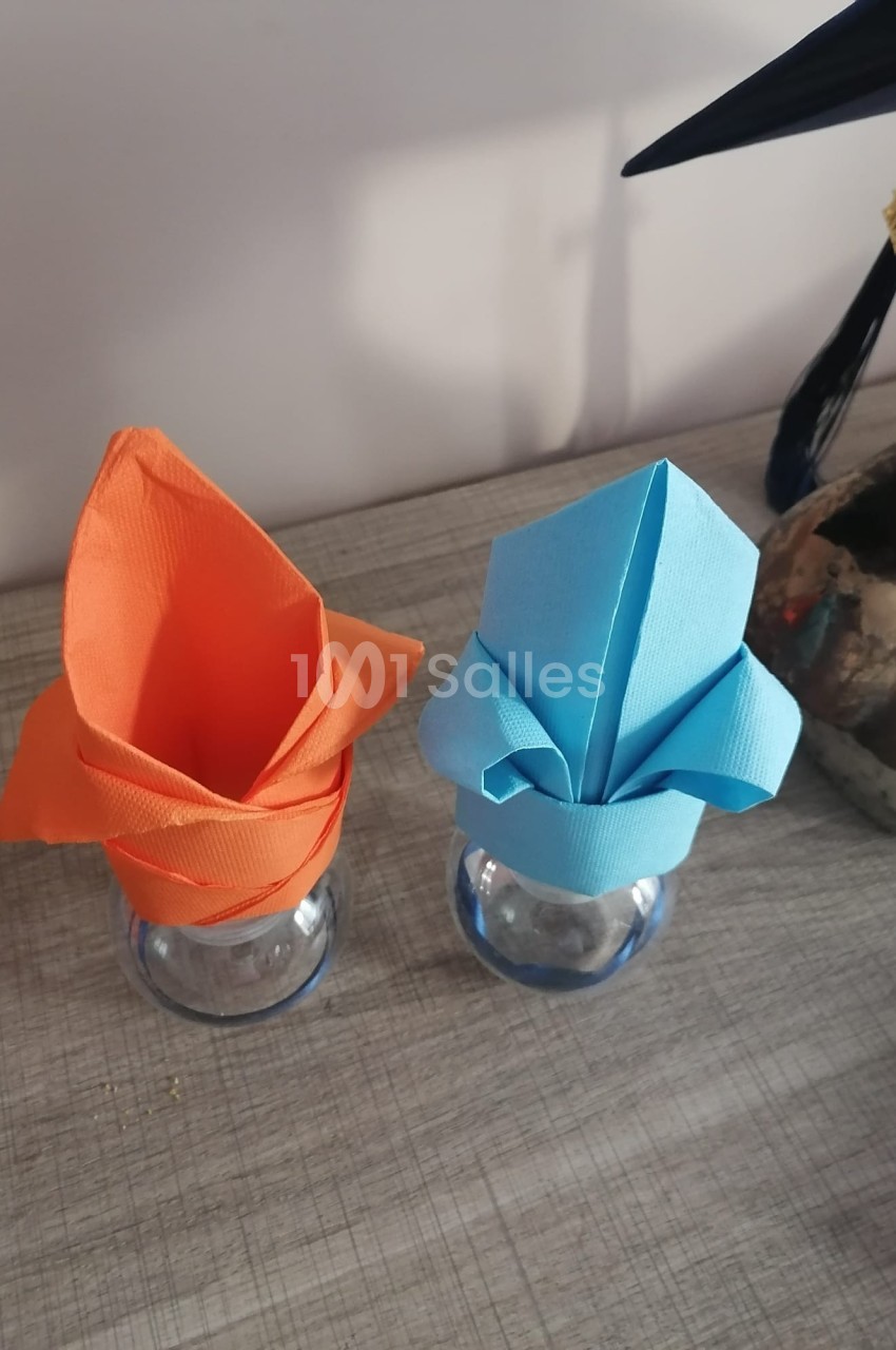Serviettes pliées en forme de fleurs, orange et bleue, disposées sur des verres transparents sur une table en bois.