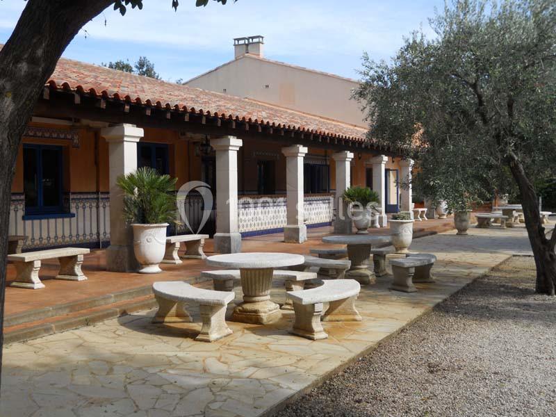 Terrasse d'une maison méditerranéenne avec tables en pierre, colonnes, et arbres environnants.