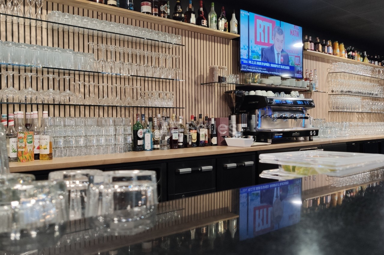 Bar moderne avec comptoir en marbre noir, étagères remplies de verres et bouteilles, machine à café et écran allumé.
