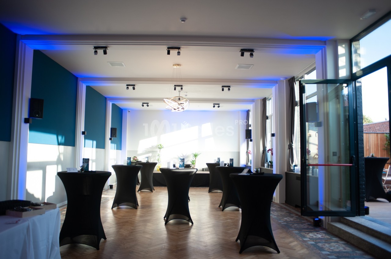 Salle lumineuse avec tables hautes noires, éclairage bleu au plafond et grandes baies vitrées ouvertes.