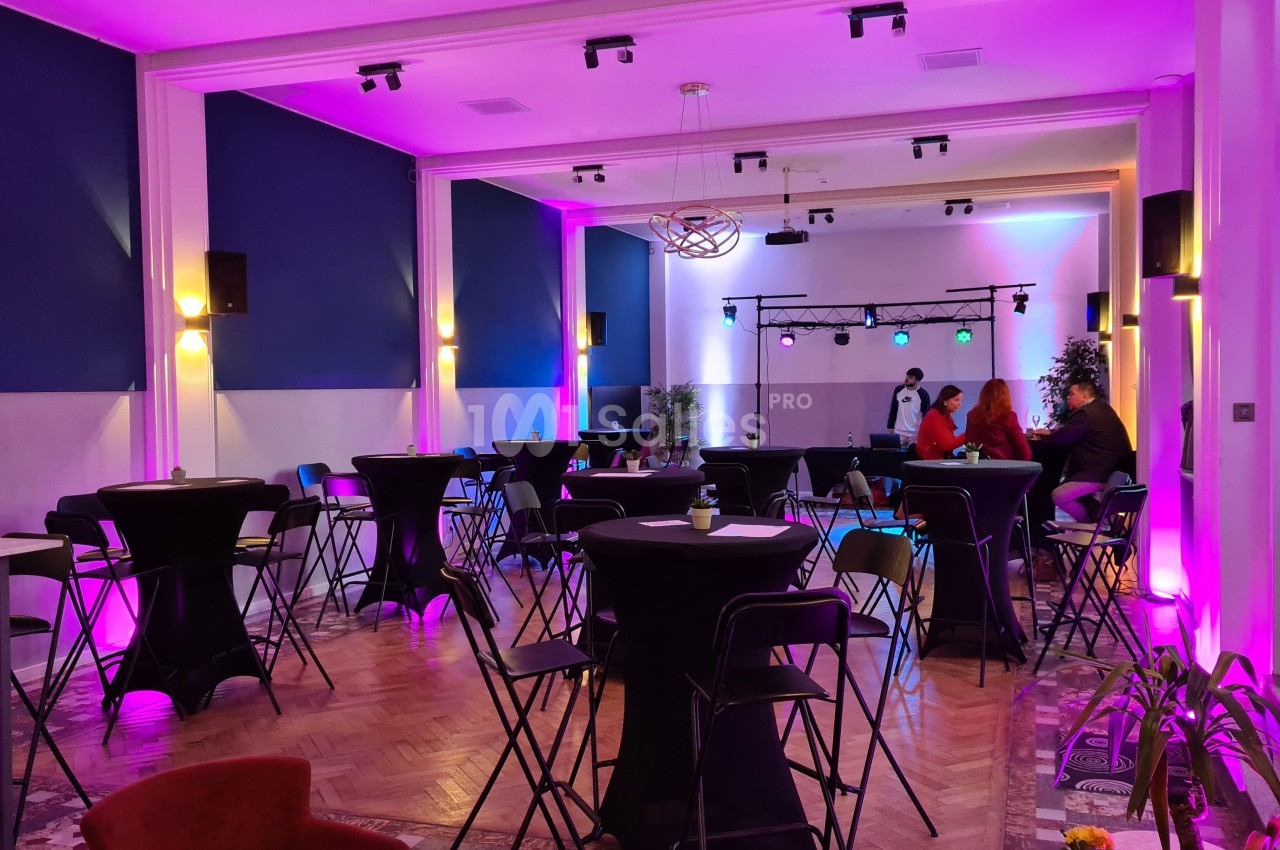 Salle de réception avec tables hautes, chaises pliantes et éclairage coloré, quelques personnes discutent au fond.