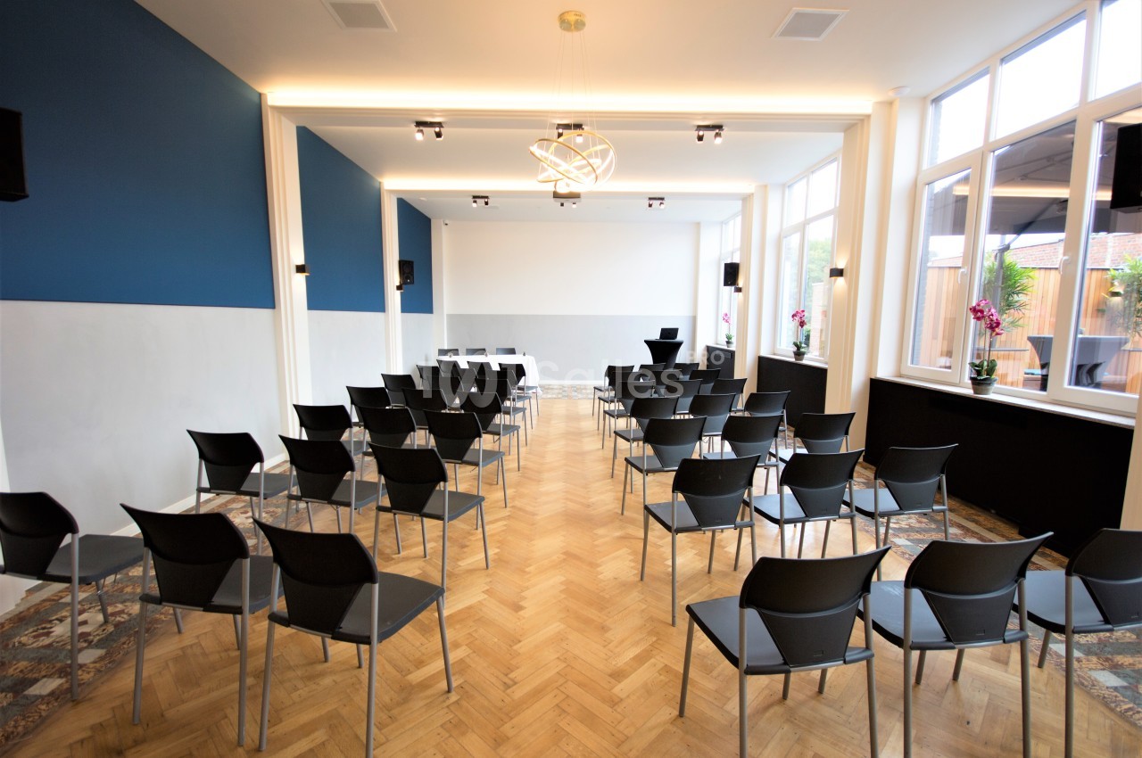 Salle de conférence lumineuse avec des chaises alignées face à un écran blanc, parquet en bois et grandes fenêtres.