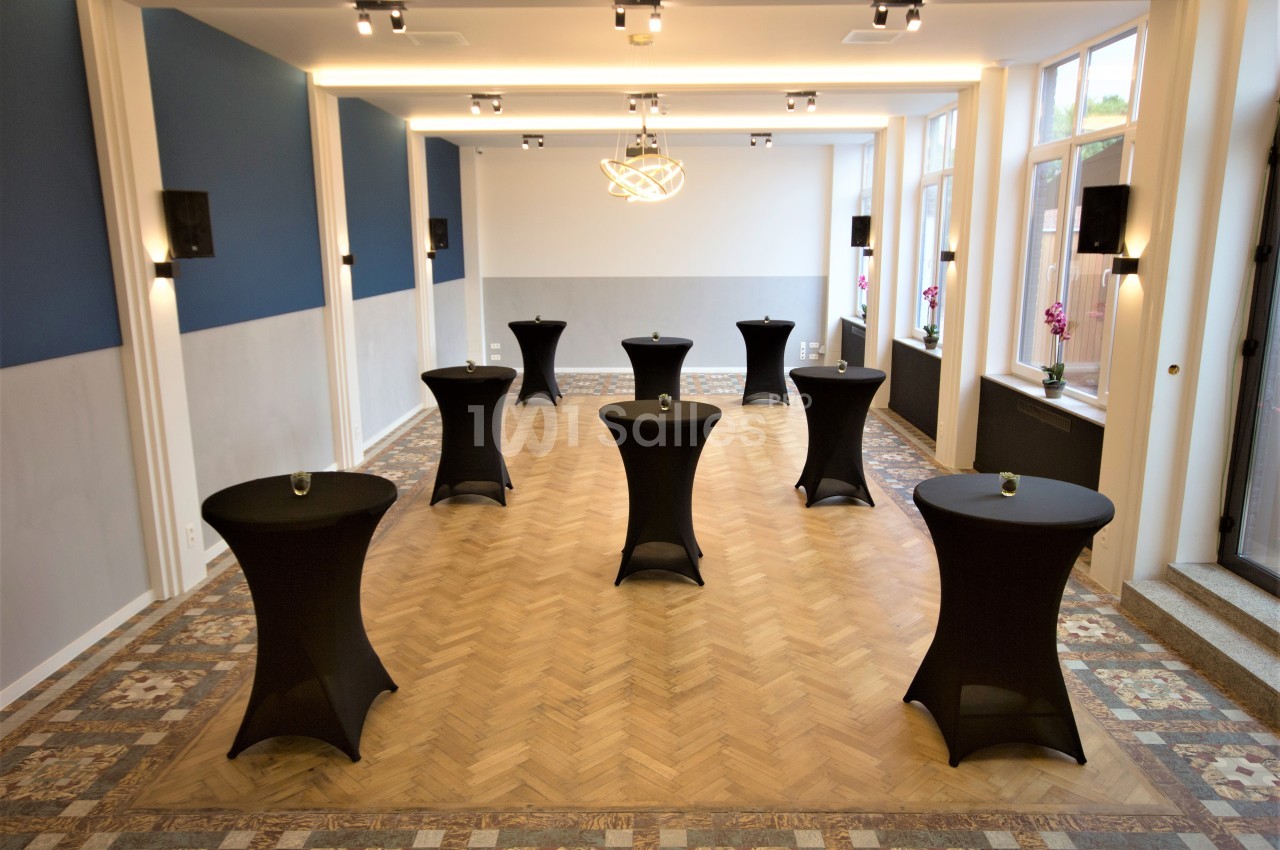 Salle lumineuse avec parquet, tables hautes noires et murs bleu et blanc, éclairée par un lustre moderne.