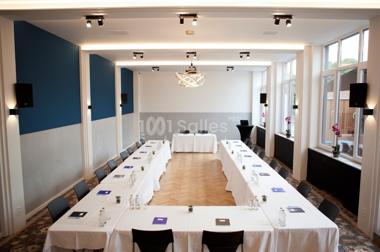Salle de réunion lumineuse avec tables disposées en U, chaises noires et documents posés sur les tables.