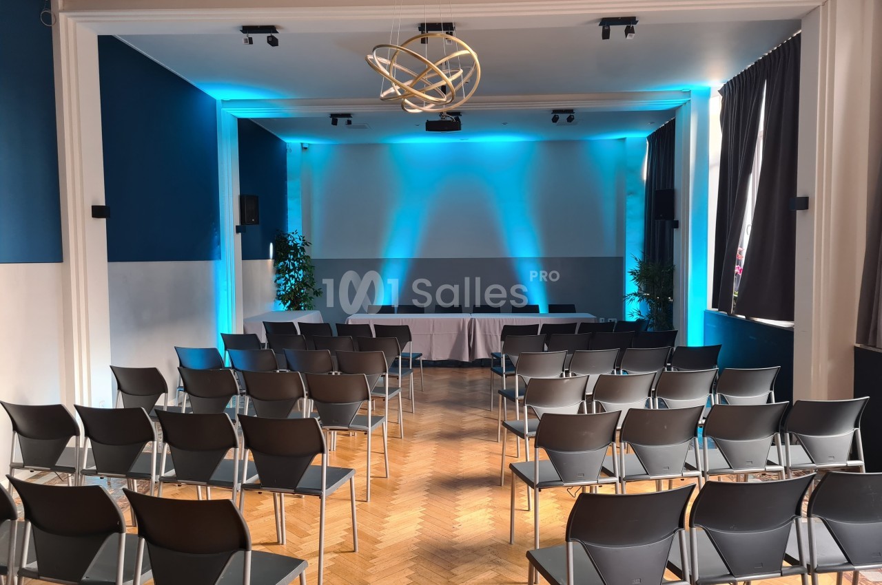 Salle de conférence avec des rangées de chaises noires, une table blanche et un éclairage bleu sur le mur du fond.