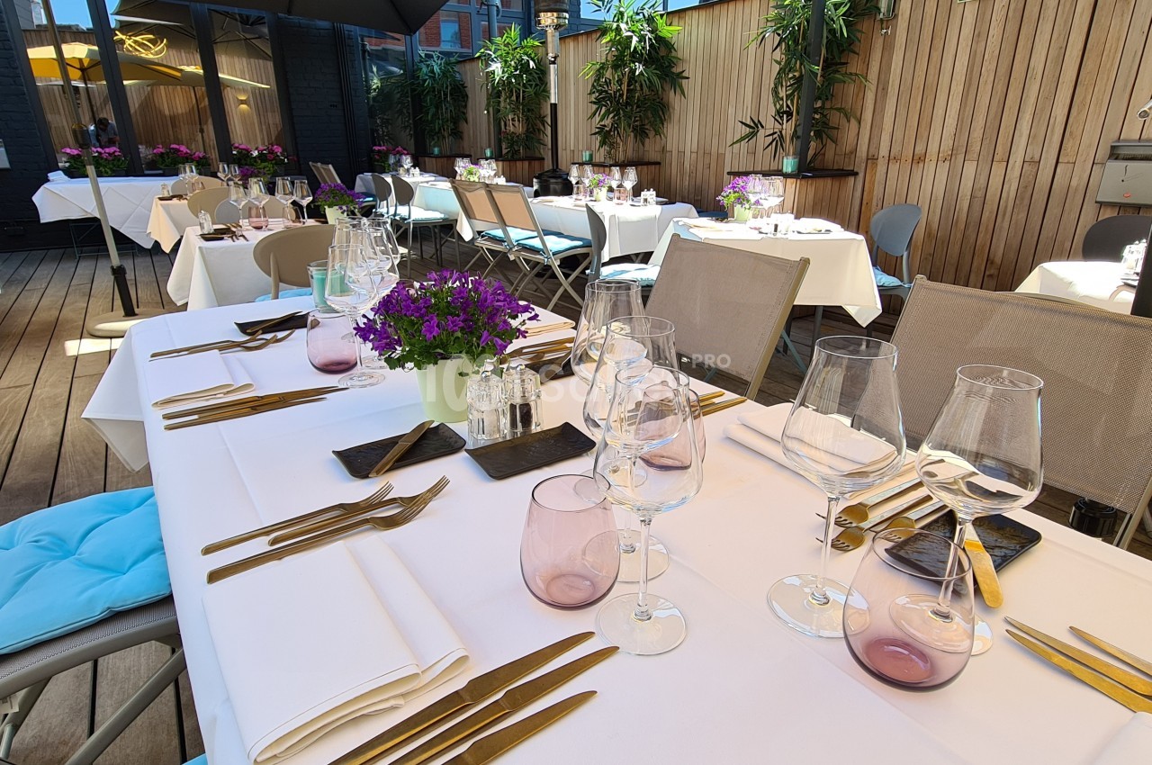 Tables dressées avec nappes blanches, verres et couverts, sur une terrasse ensoleillée entourée de plantes.