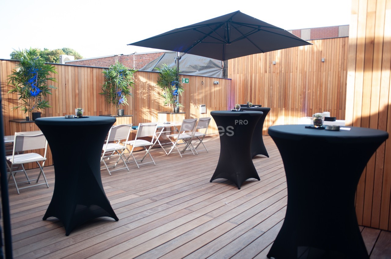 Terrasse en bois aménagée avec des tables hautes noires, des chaises blanches et des plantes en fond.