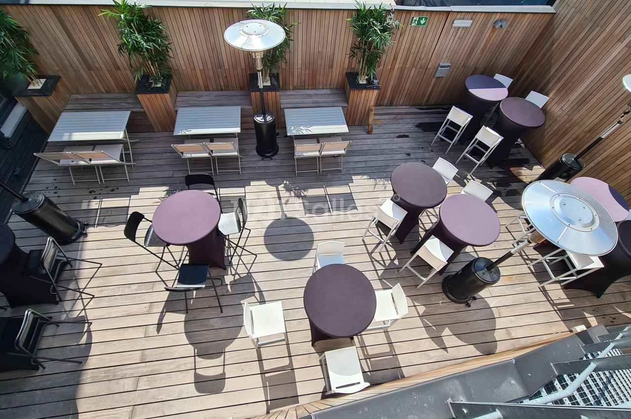 Terrasse en bois aménagée avec des tables rondes et rectangulaires, chaises blanches et chauffages extérieurs.