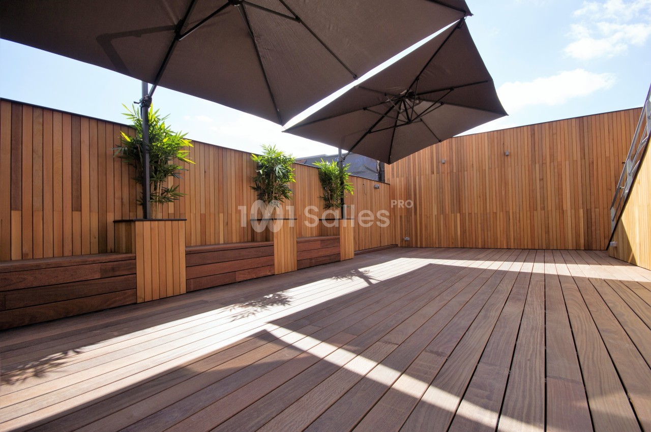 Terrasse en bois avec des parasols noirs et des jardinières contenant des plantes vertes, entourée de panneaux en bois.