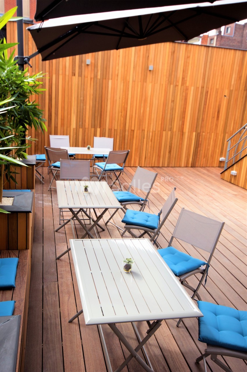 Terrasse en bois avec tables blanches, chaises pliantes à coussins bleus et plantes en pot, partiellement ombragée.