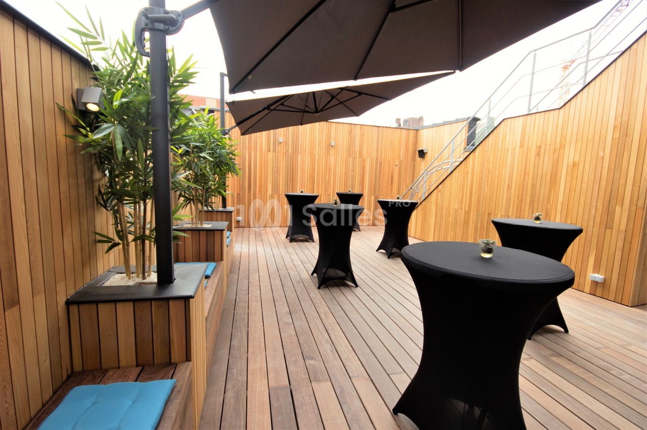 Terrasse en bois avec tables hautes couvertes de nappes noires, banquettes, plantes et parasols.