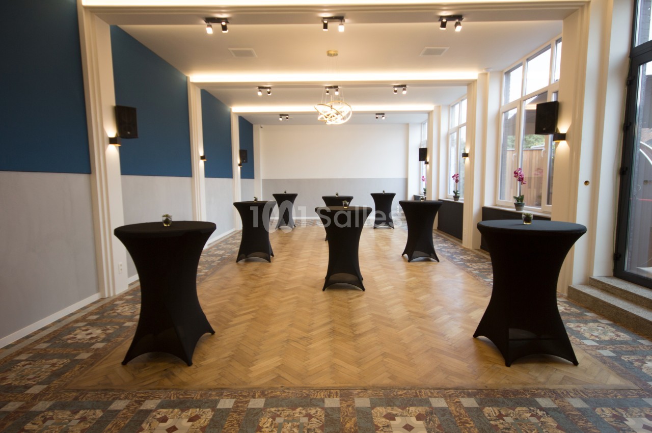 Salle lumineuse avec parquet en bois, tables hautes noires et décor moderne, adaptée pour événements ou réceptions.