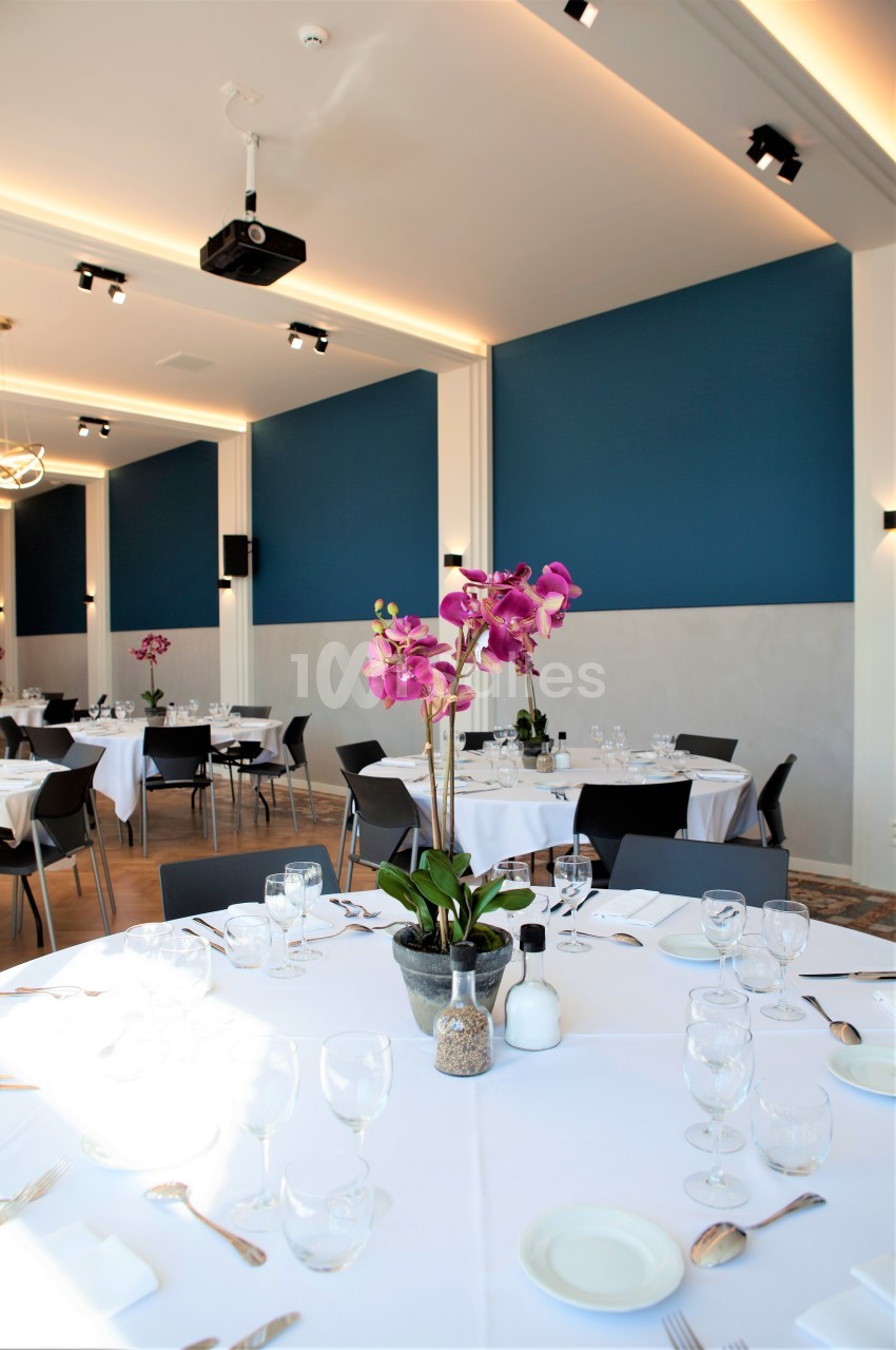 Salle de réception lumineuse avec tables rondes dressées, nappes blanches et orchidées en centre de table.