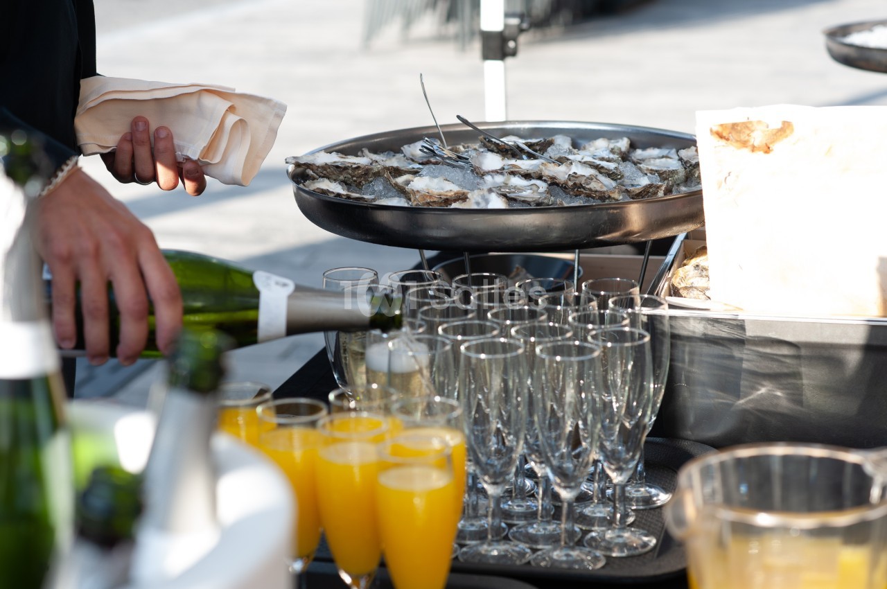 Plateau d'huîtres sur glace avec verres de champagne et jus d'orange, une main servant une bouteille de vin.