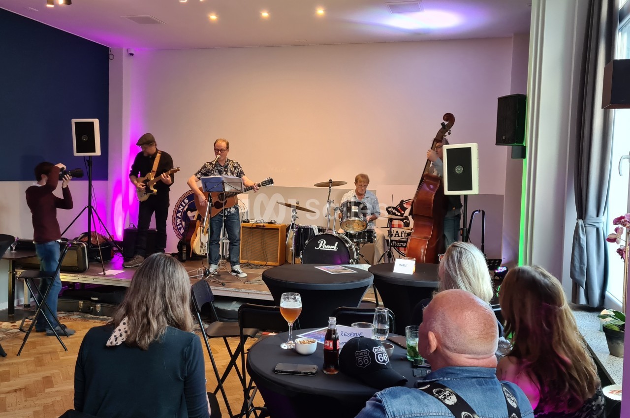 Un groupe de musiciens joue sur scène dans une salle, devant un public assis à des tables.
