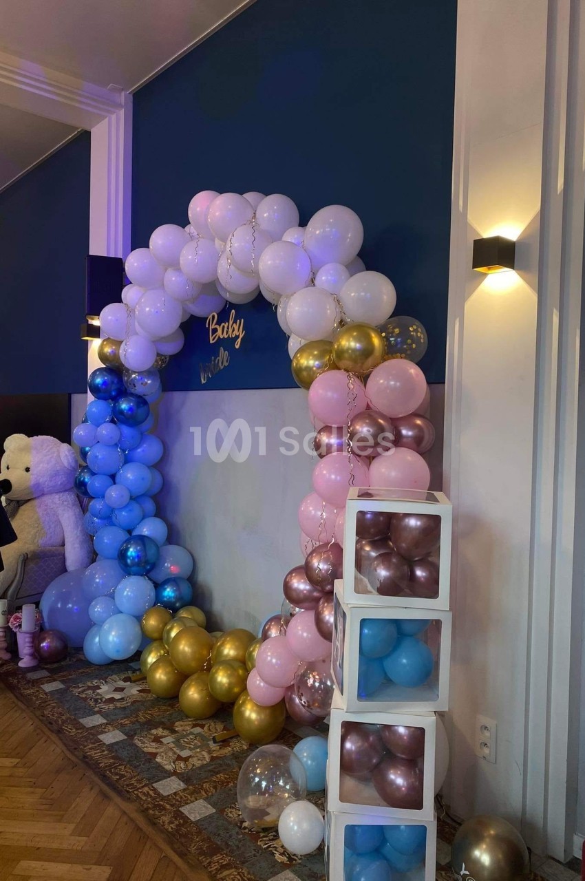 Arche décorative en ballons roses, bleus et dorés, entourée de cubes et d'un ours en peluche dans une salle éclairée.