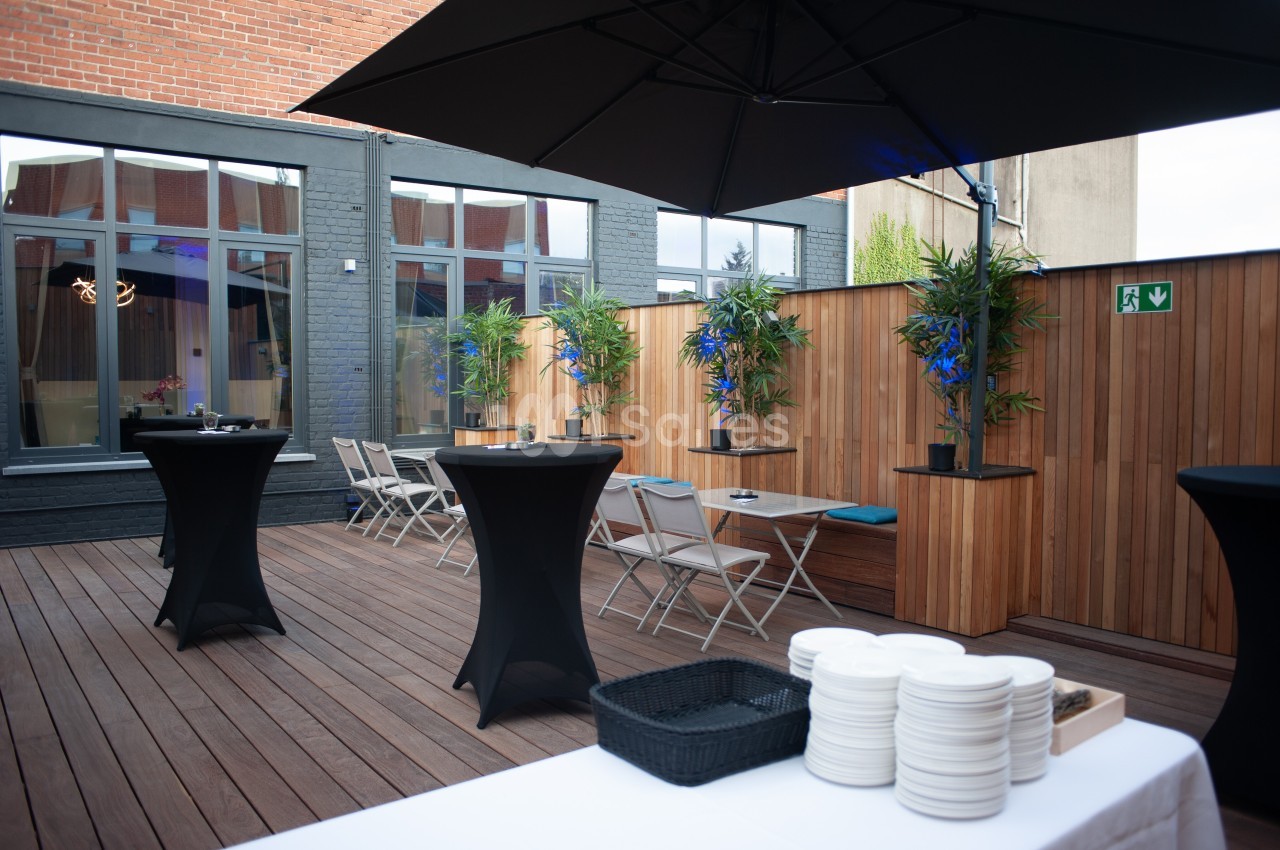 Terrasse en bois aménagée avec tables hautes, chaises pliantes, plantes et parasol, entourée de murs en bois et briques.