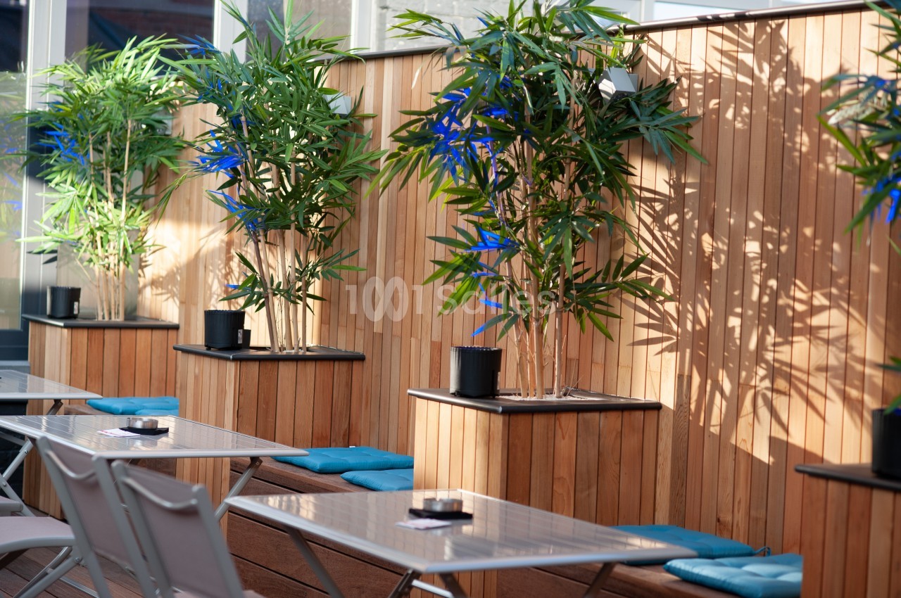 Terrasse avec tables en métal, chaises pliantes et plantes en pot devant un mur en bois éclairé par le soleil.