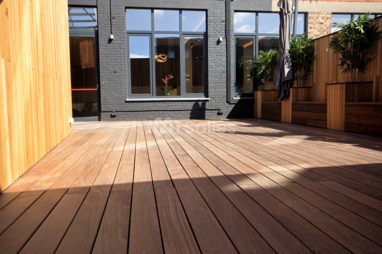 Terrasse en bois avec des murs en bois et en briques, entourée de plantes et baignée de lumière naturelle.