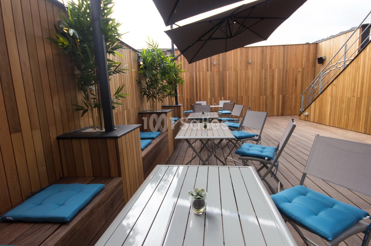 Terrasse en bois avec tables, chaises à coussins bleus, plantes et parasols, dans un espace extérieur moderne.