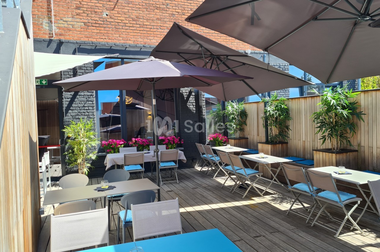Terrasse en bois avec tables, chaises, parasols et plantes, entourée de murs en briques et panneaux en bois.