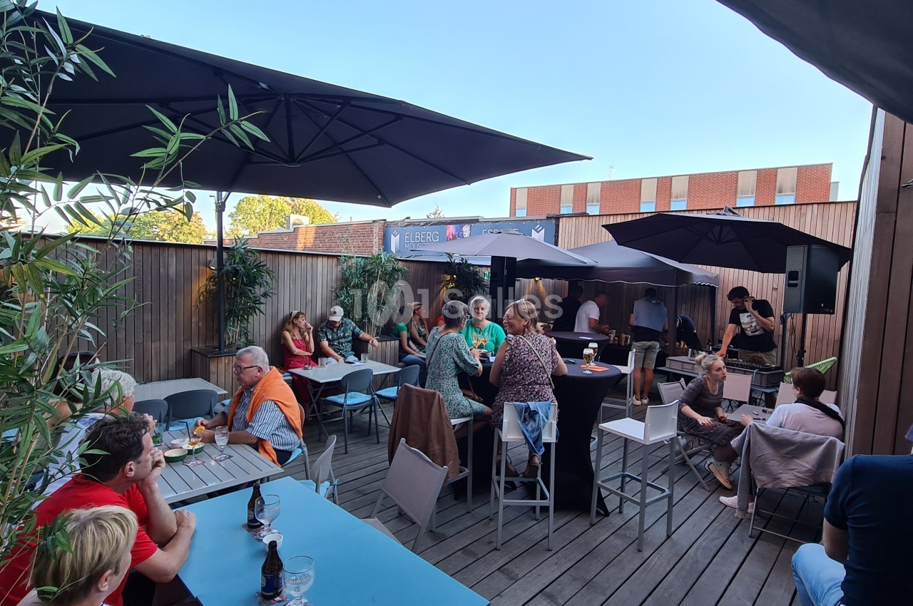 Personnes assises et debout sur une terrasse en bois, discutant autour de tables sous des parasols par temps clair.
