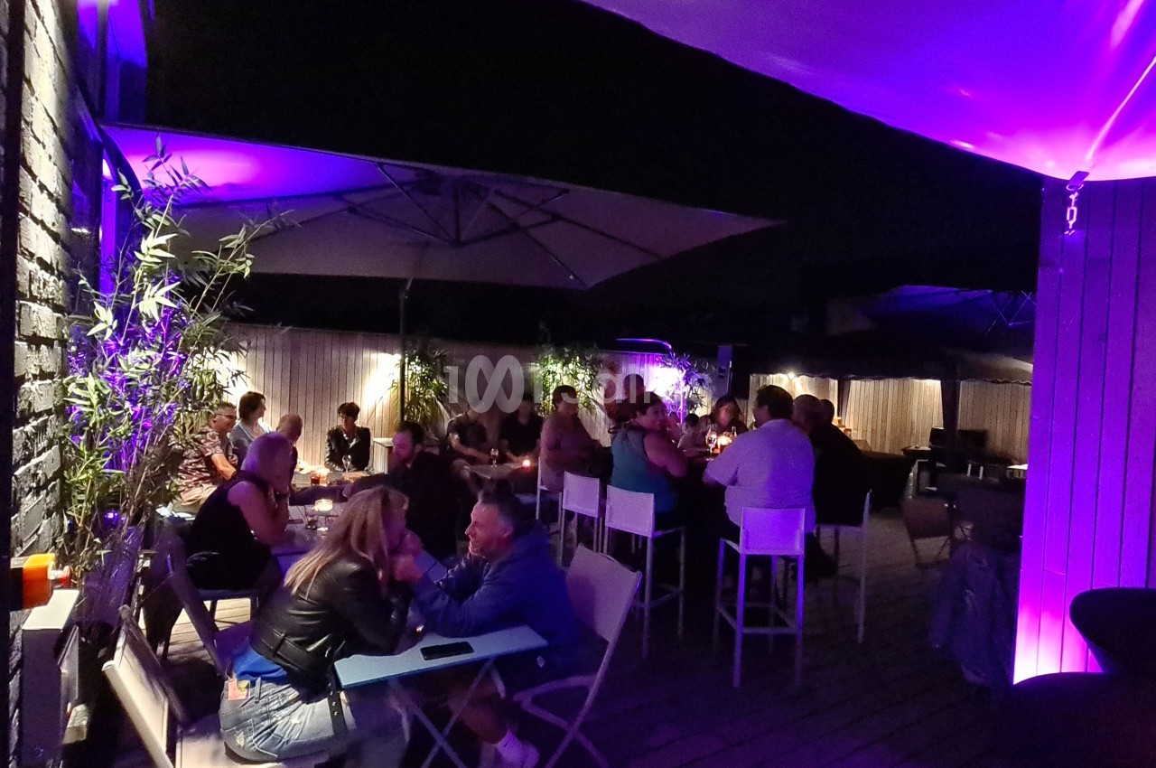 Terrasse extérieure éclairée en violet, avec des groupes de personnes assises et discutant autour de tables.