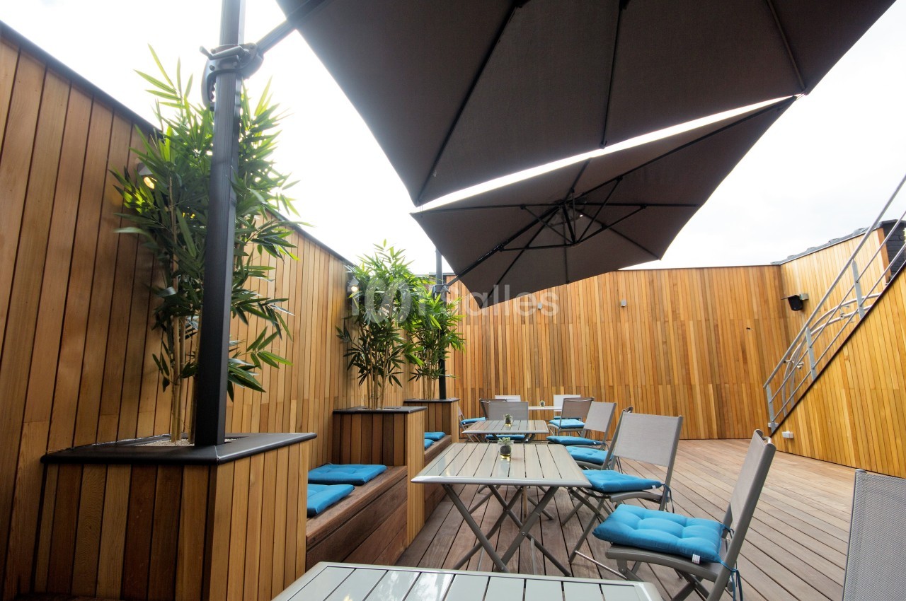 Terrasse en bois avec tables, chaises, coussins bleus et parasols noirs, entourée de panneaux en bois et de plantes.