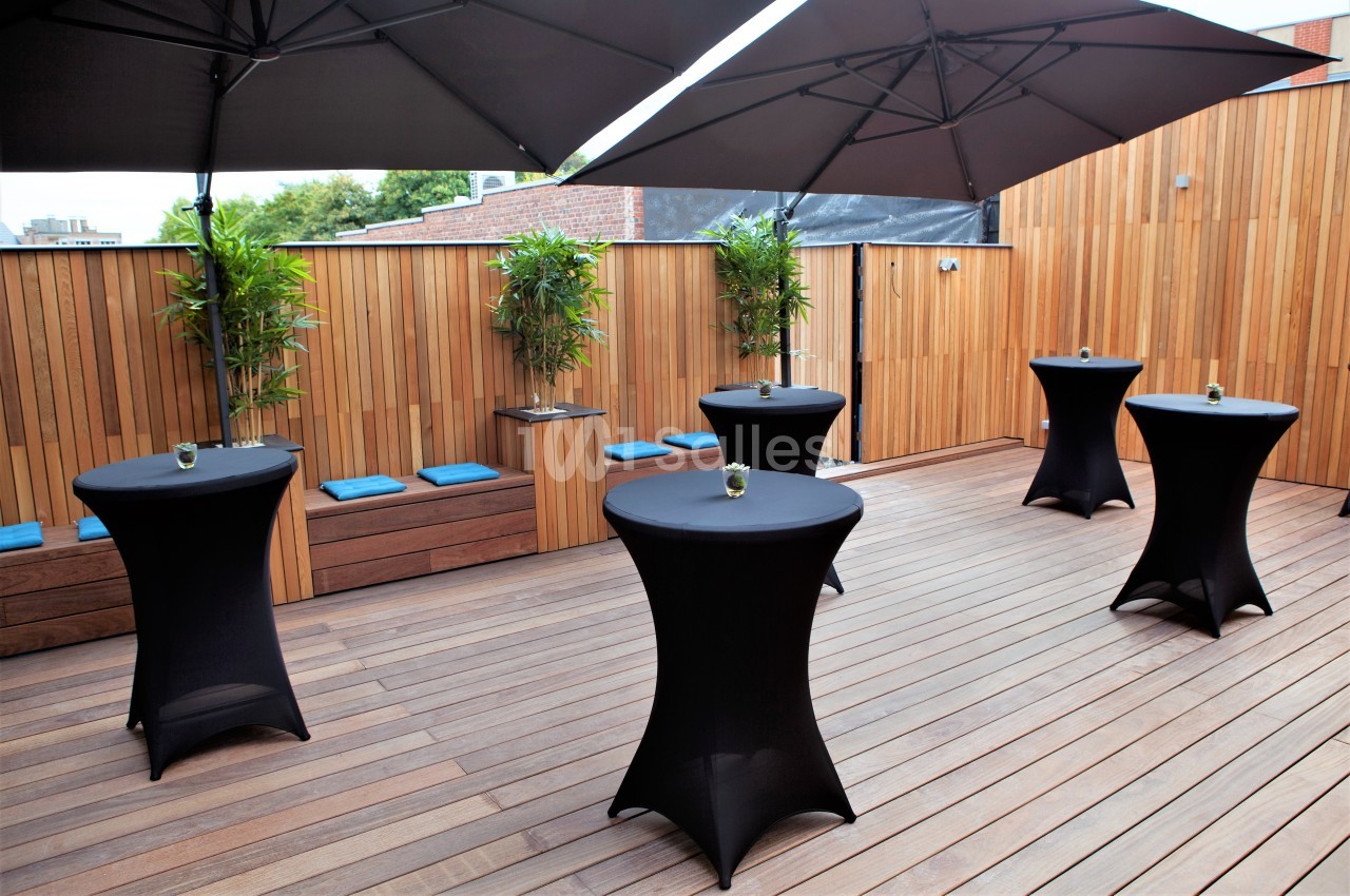 Terrasse en bois avec tables hautes noires, chaises bleues et plantes en pot, abritée par de grands parasols noirs.