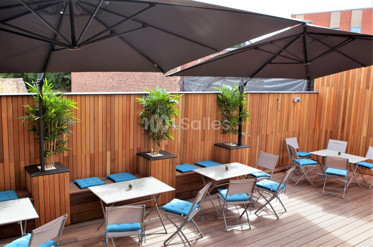 Terrasse en bois avec tables, chaises pliantes, coussins bleus, plantes en pots et grands parasols noirs.