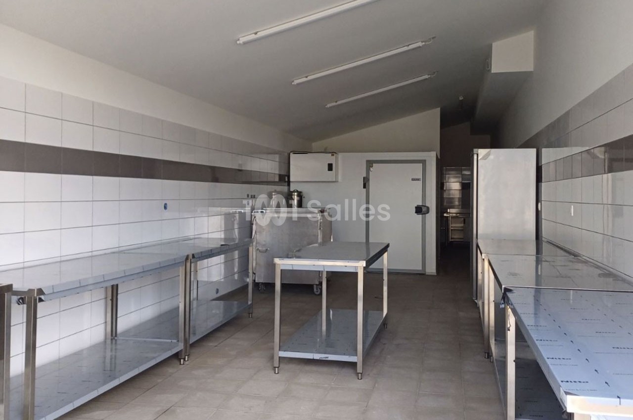 Cuisine professionnelle vide avec plans de travail en inox, murs carrelés blancs et éclairage fluorescent au plafond.