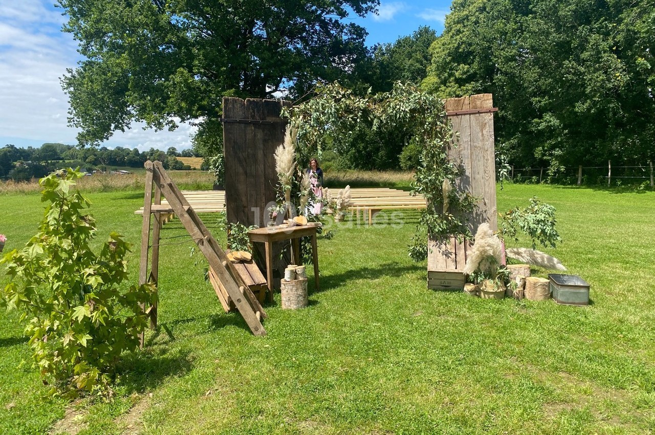 Décor champêtre avec arche en bois ornée de feuillage, bancs en arrière-plan sur une pelouse entourée d'arbres.
