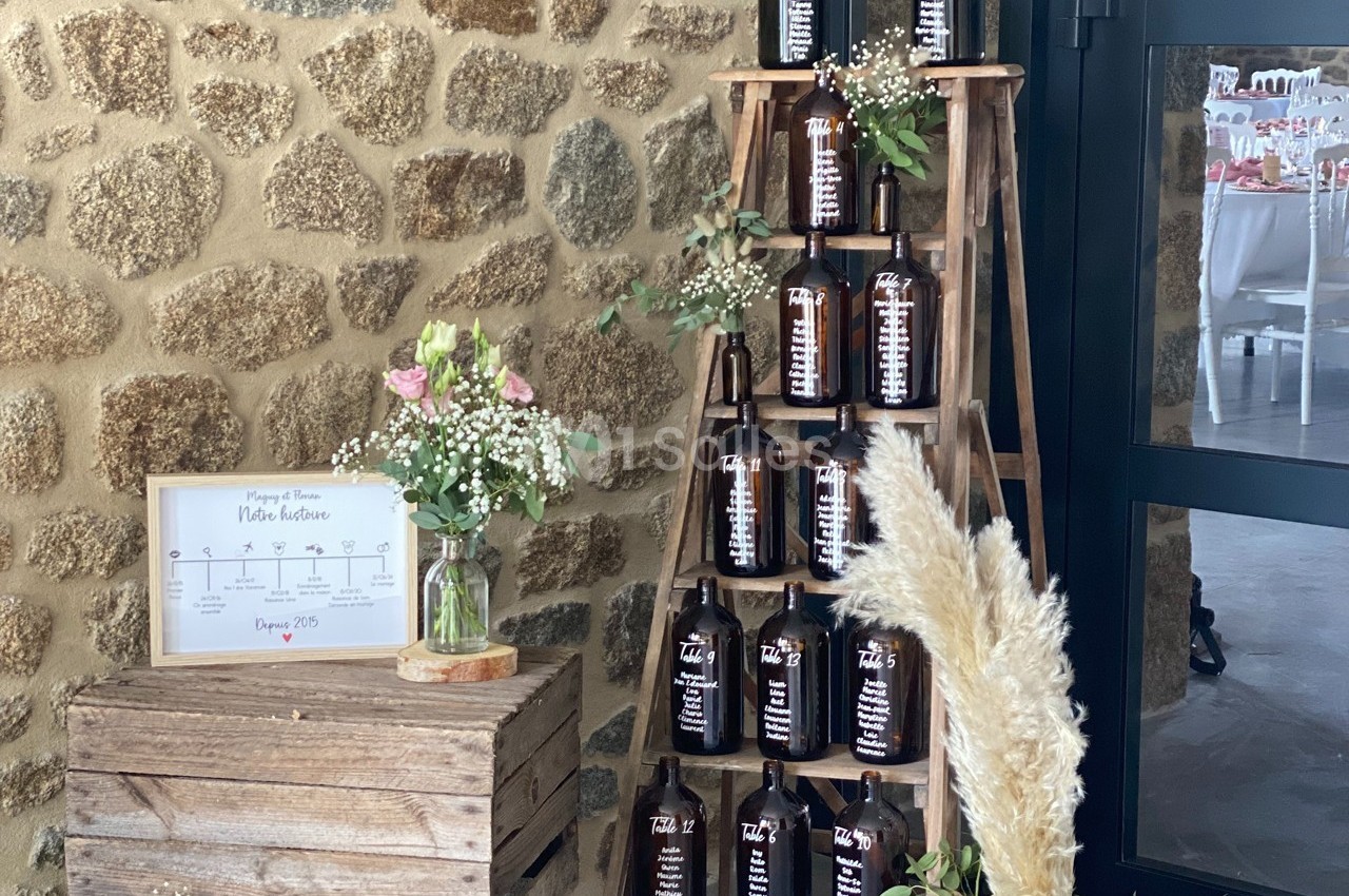 Échelle en bois décorée de bouteilles en verre marron, fleurs séchées et cadre sur fond de mur en pierre.