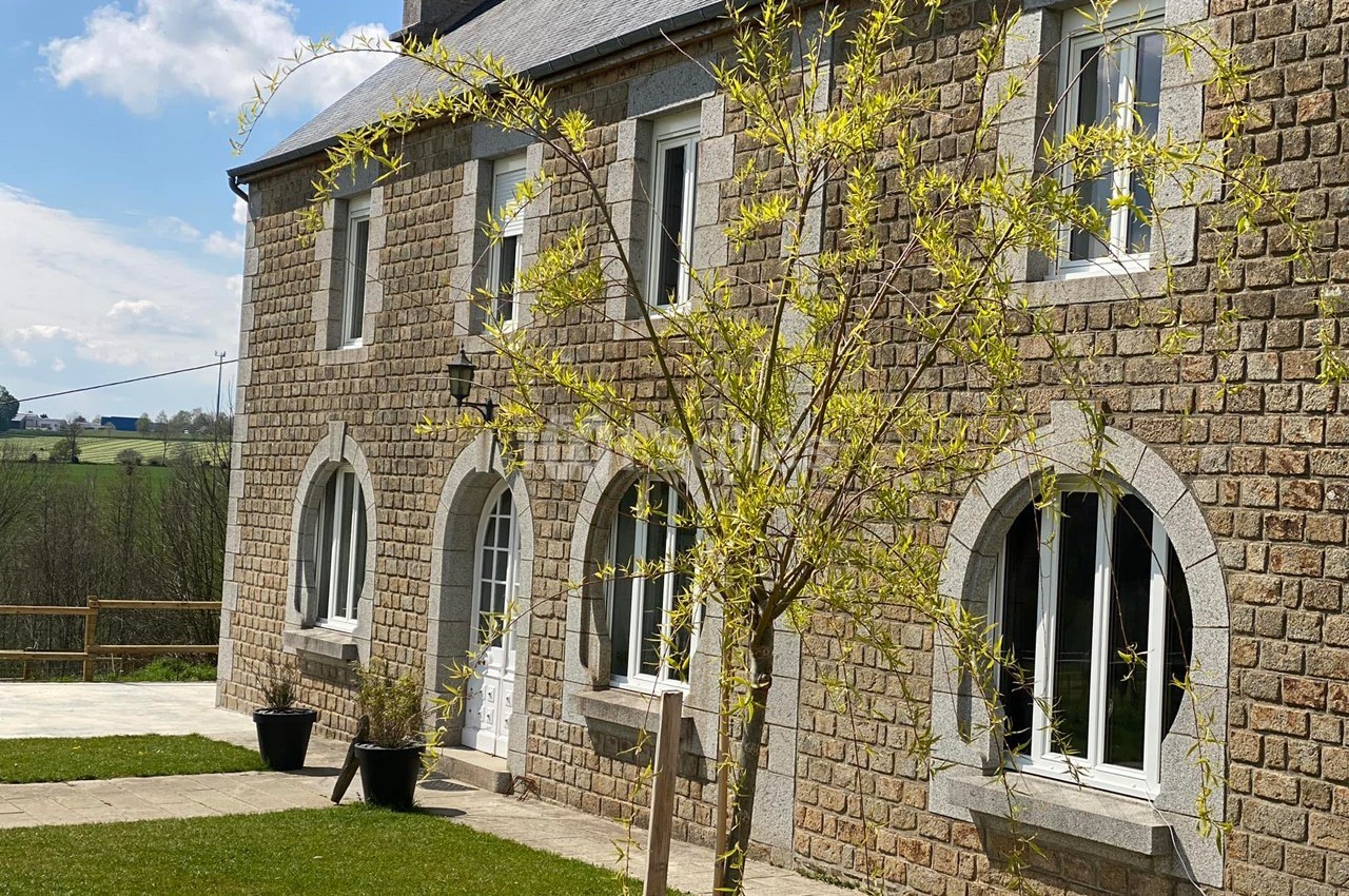 Façade en pierre d'une maison avec fenêtres cintrées, arbre jeune au premier plan et paysage rural en arrière-plan.