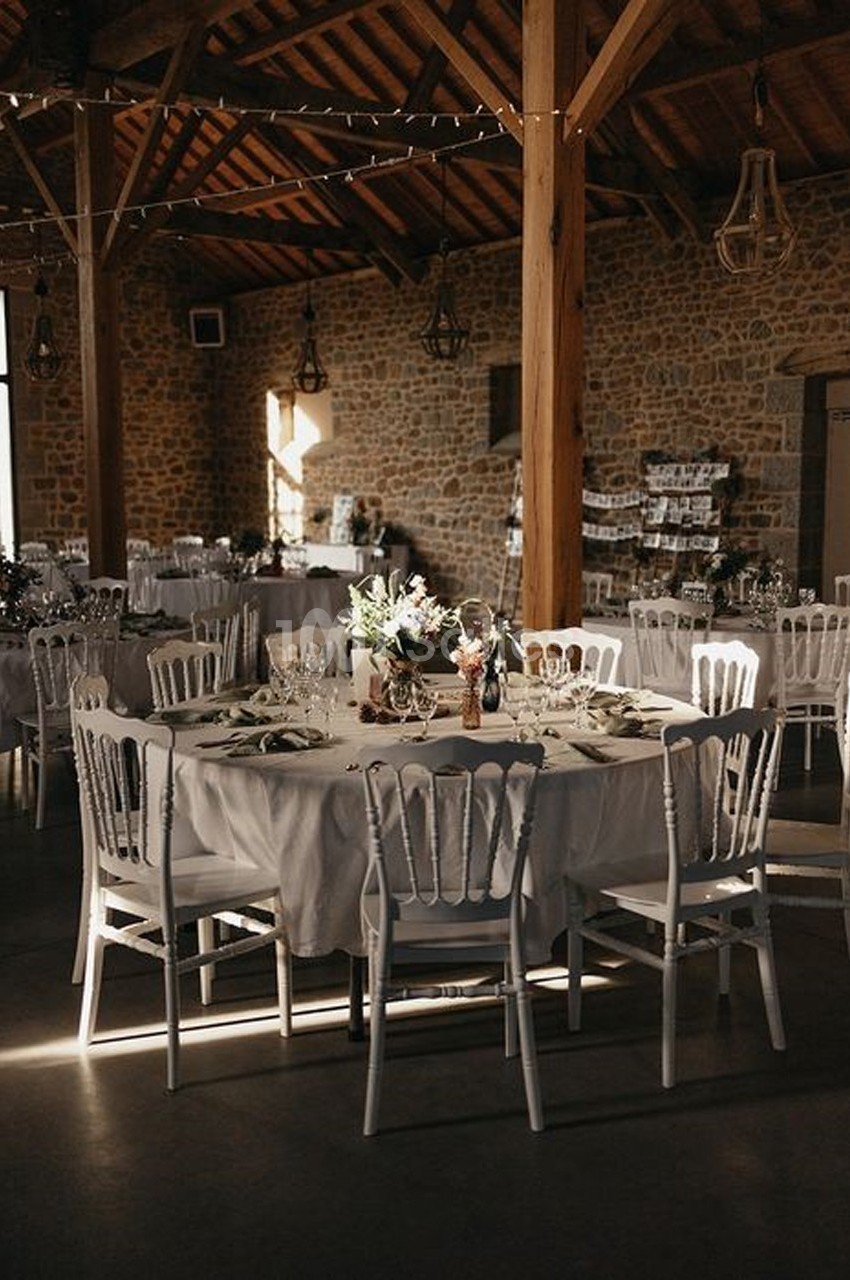 Salle de réception en pierre avec tables rondes dressées, nappes blanches et chaises blanches, éclairée par une lumière…